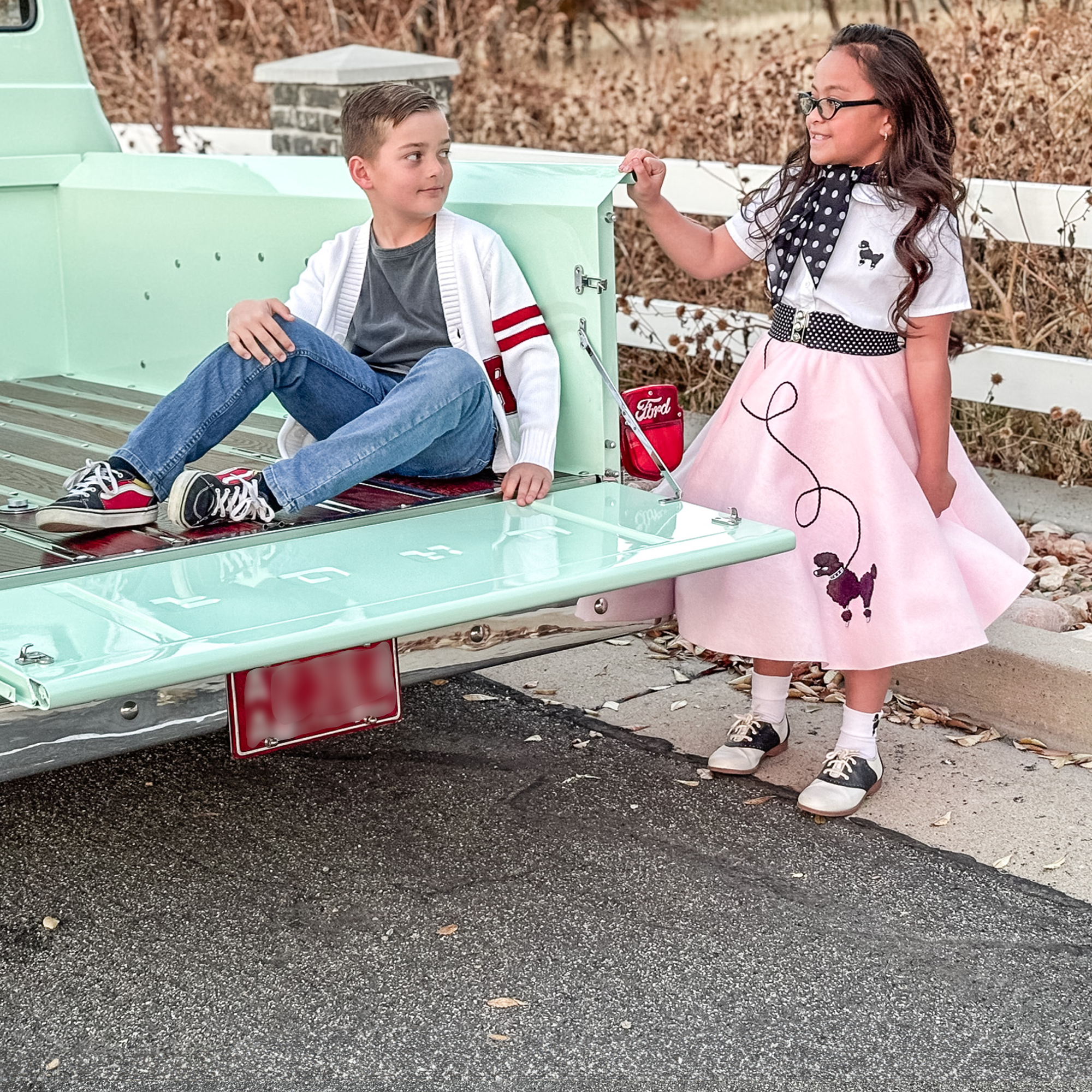Boy and Girl sitting in the back of a 1956 mint colored Ford Truck F100 modeling a retro letterman sweater and poodle skirt outfit.