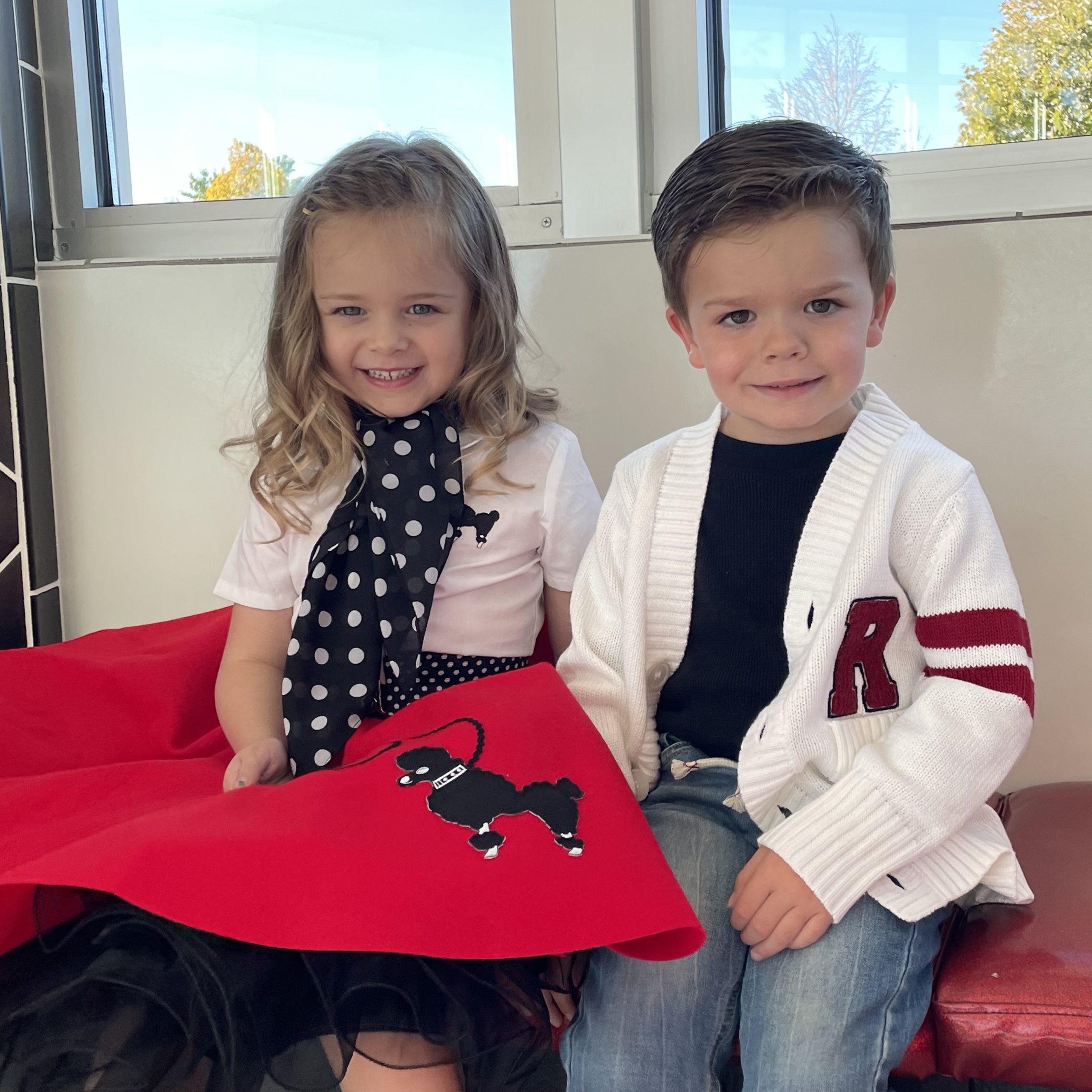 Friends sitting in an ice cream shop - toddler girl wearing a red poodle skirt outfit with polka dot accessories and a toddler boy wearing a white retro letterman sweater with an R monogram over the left pocket with stripes on the sleeve.