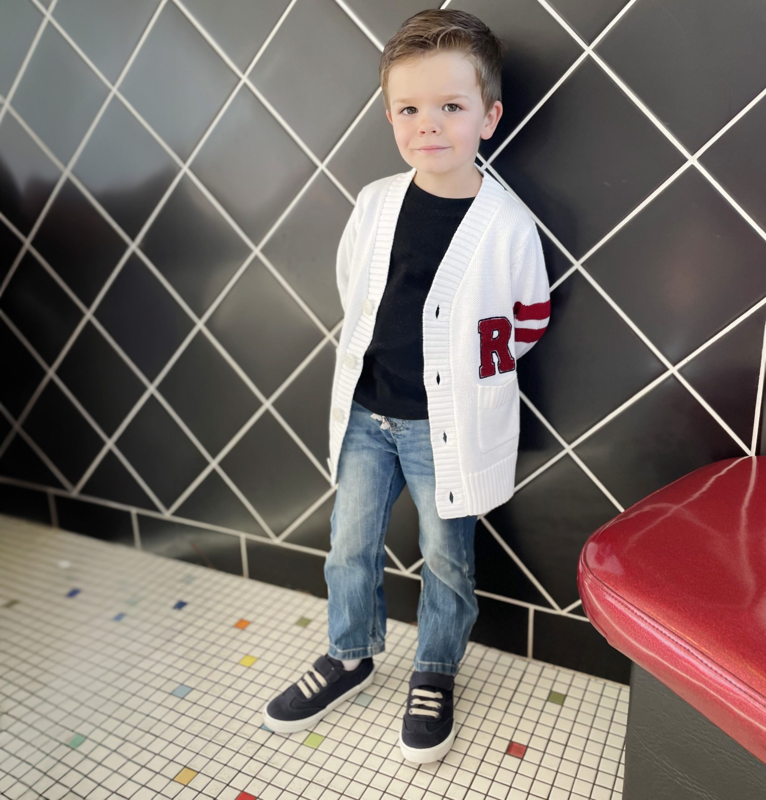 Toddler boy modeling a retro letterman sweater with an R monogram over the left pocket with two stripes on sleeve in a diner.